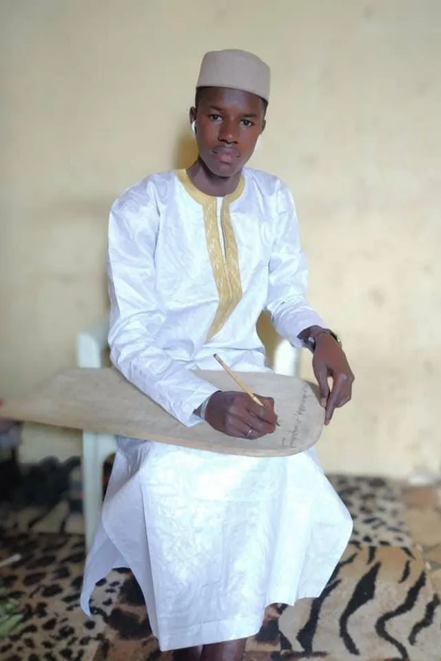 Mouhamed Nour All-Haqq en boubou blanc, écrivant au calame sur une planchette coranique en bois